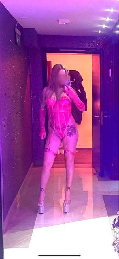 Eine Person in einem pinken Korsett-Set mit Strapsen und High Heels hält ein Smartphone. Im Hintergrund eine Tür mit Kette und zarte Lichter an der Wand.