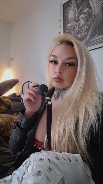Eine Person mit blonden Haaren, Tattoos am Hals und Hand, trägt ein schwarzes Lederoutfit und hält ein Mikrofon. Im Hintergrund ein Gemälde und ein Kamin.