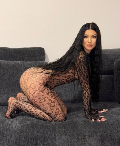 Eine Person mit langen schwarzen Haaren kniet auf einem grauen Sofa. Sie trägt ein hautenges, durchsichtiges Outfit mit Schlangenmuster und hat auffällige Nägel.