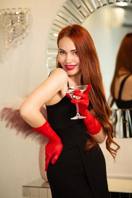 Eine Frau mit roten Handschuhen und einem schwarzen Kleid hält ein Cocktailglas. Im Hintergrund sind ein Spiegel, ein Schmuckstück und ein Gefäß mit Federn zu sehen.
