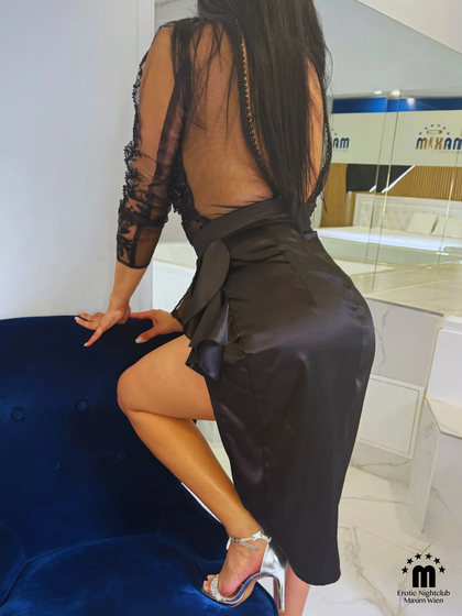 Eine Frau in einem schwarzen, durchsichtigen Kleid mit Rückschnitt steht mit dem Rücken zur Kamera. Sie stützt sich auf einen blauen Sessel, trägt silberne High Heels und hat langes, dunkles Haar.