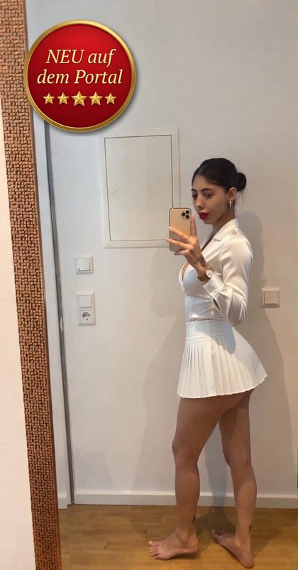 Eine Frau in weißem Outfit macht ein Selfie im Spiegel. Sie trägt einen Blazer und einen kurzen Rock. Im Hintergrund eine weiße Tür. Oben links ein rotes Emblem mit "NEU auf dem Portal".