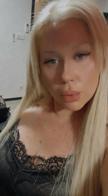 Eine Frau mit langem blonden Haar, leichten make-up und grünlichen Augen trägt ein schwarzes Spitzenoberteil. Hinter ihr ist eine Wand mit einem Schalter und einem Steckdose zu sehen.