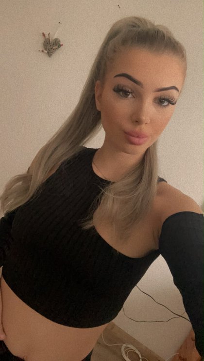 EinBLondine mit langen Haaren und perfektem Make-up trägt ein schwarzes, Schulterfreies Oberteil und hält im Raum Musikdraht in der Hand.