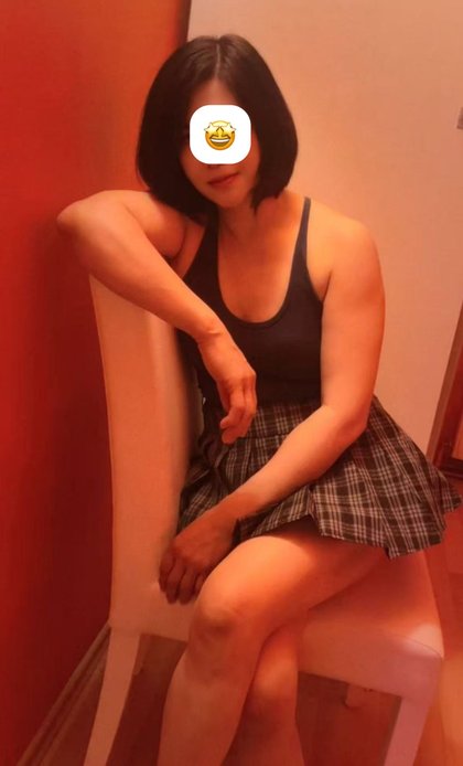 Eine Person mit kurzen, dunklen Haaren sitzt auf einem Geländer. Sie trägt ein schwarzes Top und einen karierten Rock. Ein Emoji bedeckt das Gesicht.