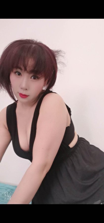 Eine Person mit kurzen, lila gefärbten Haaren trägt ein schwarzes Kleid mit tiefem Ausschnitt und sitzt seitlich zur Kamera. Hintergrund ist weiß.