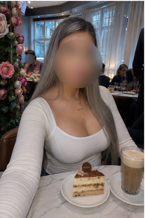Eine Person in einem weißen Top und langen Ärmeln sitzt an einem Tisch mit einem Stück Kuchen und einem Getränk. Im Hintergrund: gedämpftes Ambiente mit Kerzenlicht und Rosen.