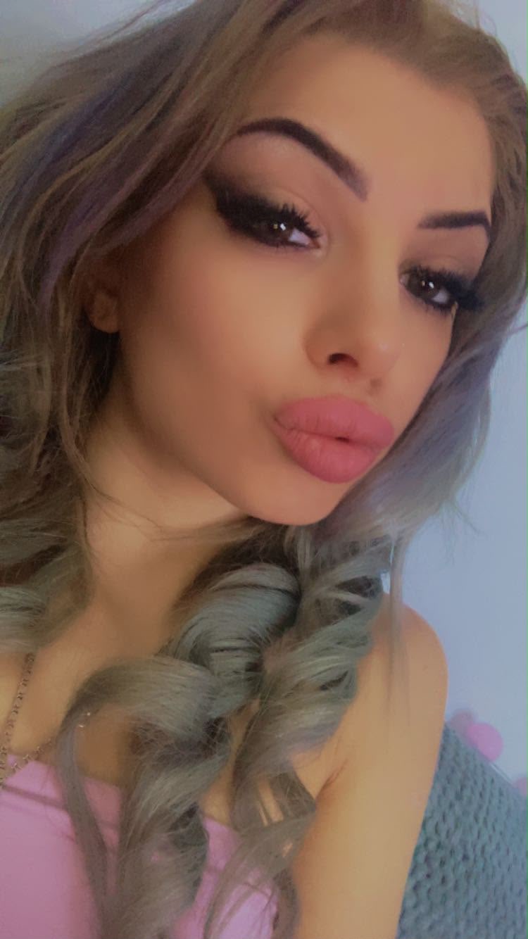 Eine Person mit gewellten, hellbraunen Haaren und auffallendem Make-up zeigt eine Zopf-Frisur. Im Hintergrund: Pastellfarben.