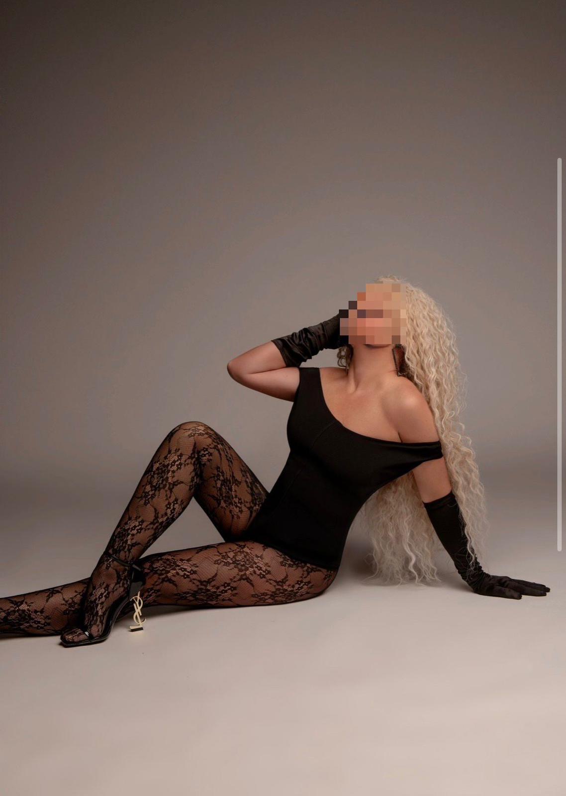 Eine sitzende Frau mit blonden Locken und geschwungener Haarsträhne trägt schwarzes Outfit mit Spitzenstrümpfen und High Heels.