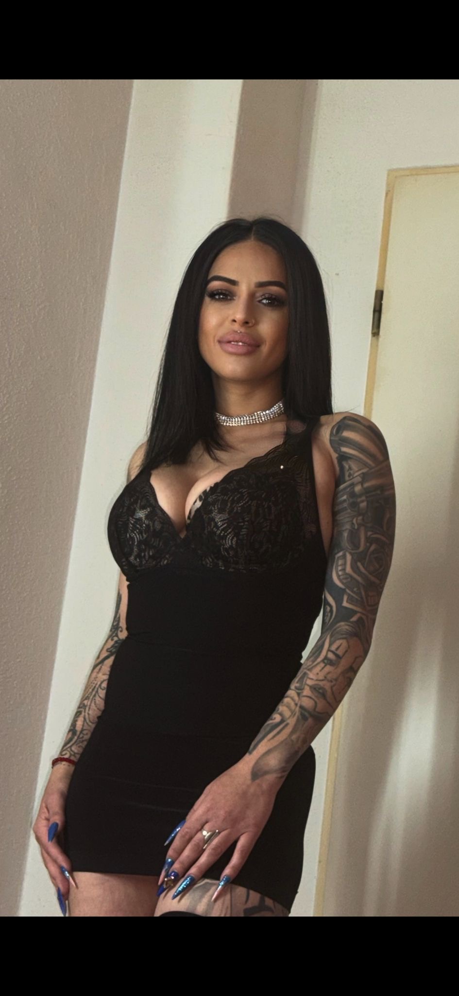 Eine Frau mit schwarzen Haaren trägt ein schwarzes Spitzenkleid und hat Tattoos an den Armen. Sie steht vor einer weißen Wand.