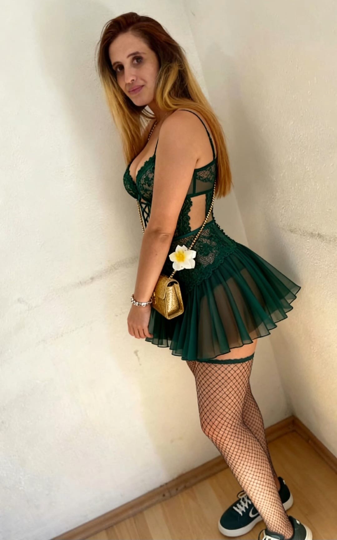 Eine Frau in einem grünen Spitzenkleid mit Netzstrümpfen und Sneakern steht an einer weißen Wand und hält eine goldene Tasche mit Blume.