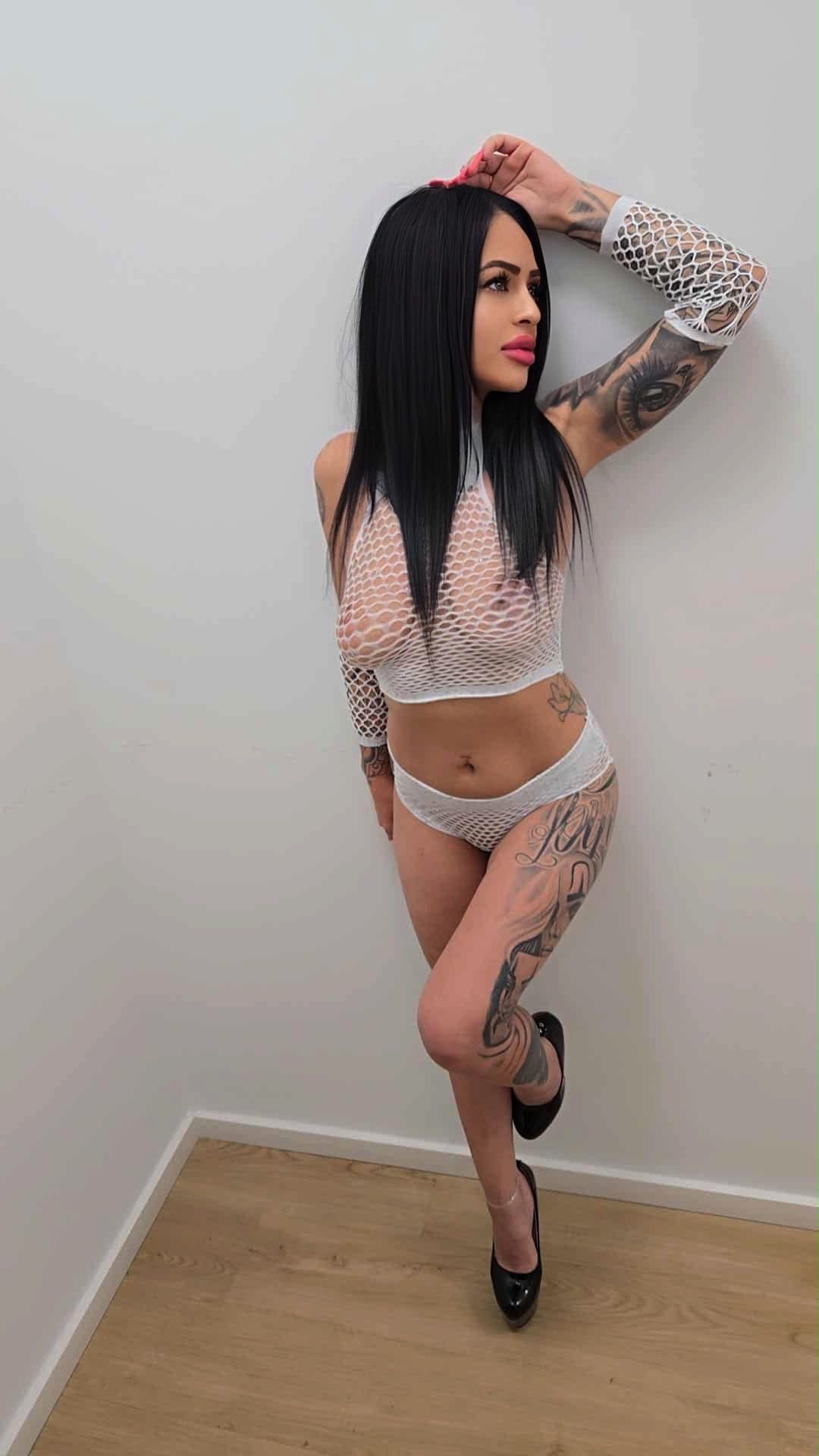 Eine Frau mit Tattoos trägt ein weißes Netz-Outfit und schwarze High Heels. Sie steht an einer weißen Wand in einem Raum mit Holzboden.