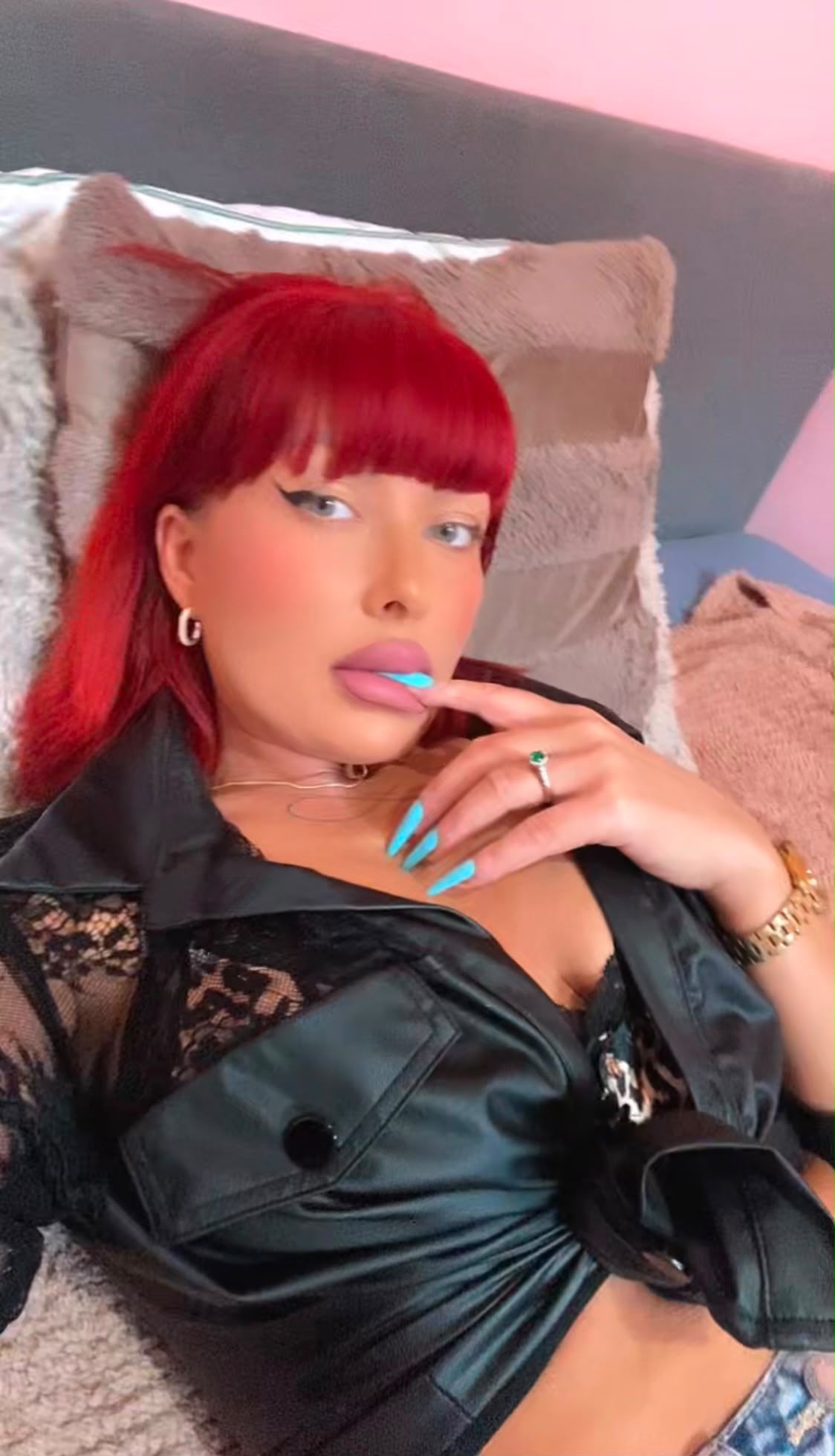 Eine Person mit roten Haaren in schwarzer, samter Jacke und Spitzen-BH liegend, Hand am Mund, Nagellack und Ring sichtbar.