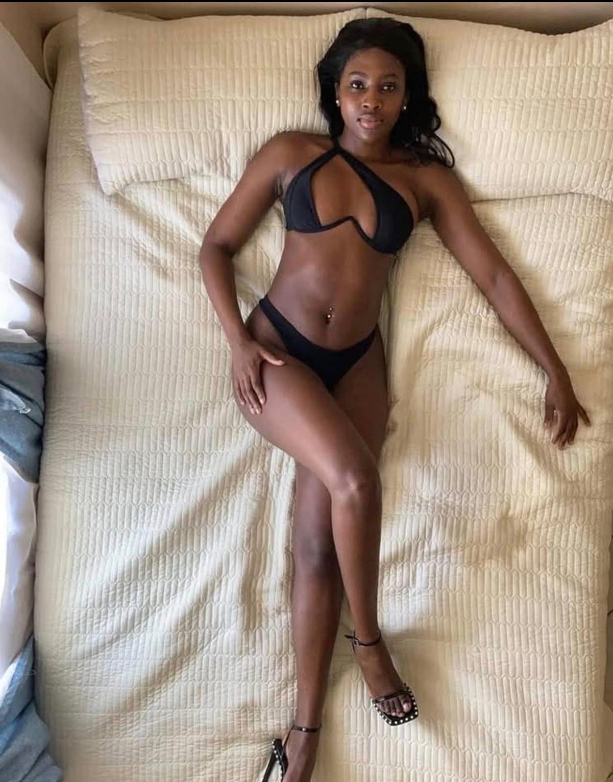 Eine Frau liegt in schwarzem Bikini auf einer hellen, strukturierten Decke. Sie trägt Sandalen und legt lässig ein Bein über das andere, während sie lächelt.
