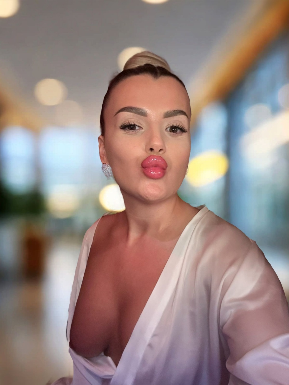 Eine Frau mit blondem Haarknoten trägt ein weißes Oberteil mit tiefem Ausschnitt und silberne Ohrringe. Sie posiert für das Bild.