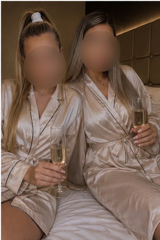 Zwei Personen in seidenen Bademänteln sitzen auf einem Bett mit Champagnergläsern in den Händen. Ihre Gesichter sind unscharf.
