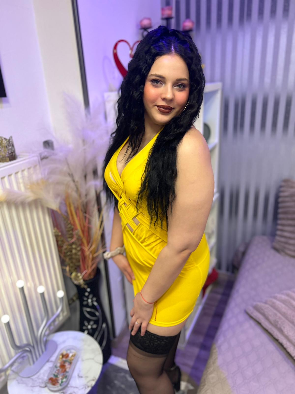 Eine Frau in einem gelben Kleid steht in einem Raum mit lila Beleuchtung. Sie trägt schwarze Strümpfe und hat langes, lockiges Haar. Im Hintergrund sind dekorative Elemente zu sehen.
