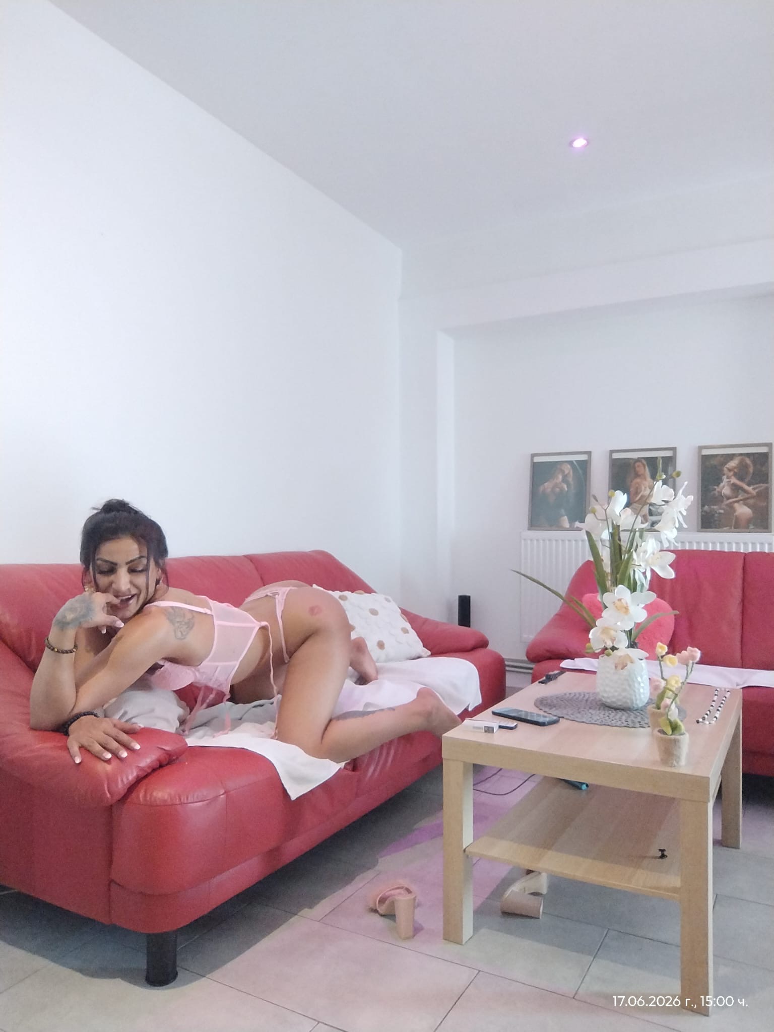 Eine lächelnde Frau in rosa Dessous liegt auf einer roten Couch im Wohnzimmer. Auf dem Couchtisch daneben stehen weiße Orchideen.