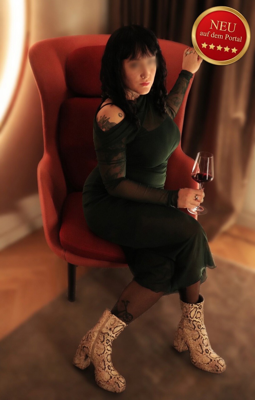 Eine Frau in einem grünen Kleid sitzt auf einem roten Sessel, hält ein Weinglas und trägt Schlangenlederstiefel. Im Hintergrund ein Vorhang.