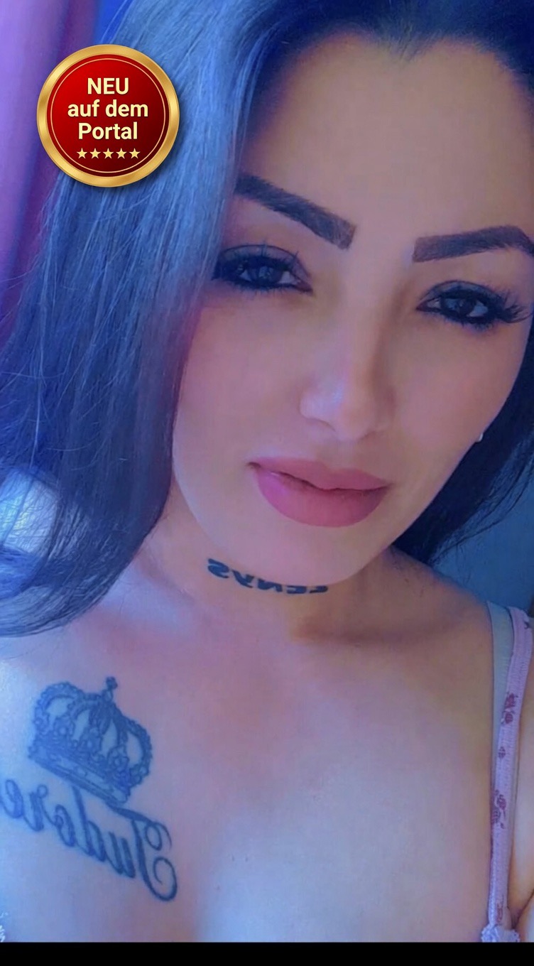 Eine Frau mit blauem Haar, geschminktem Gesicht und Tattoos am Hals und Brust. Sie trägt ein rosa Top. Ein rotes Emblem mit "NEU auf dem Portal" ist oben links.