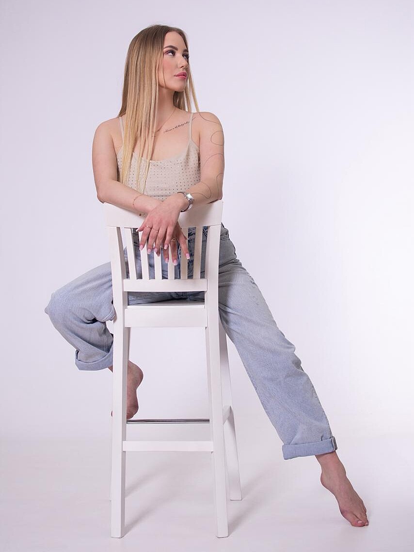 Eine junge Frau mit blonden Haaren sitzt barfuß auf einem weißen Stuhl. Sie trägt ein ärmelloses Hemd und weite Jeans. Der Hintergrund ist weiß.