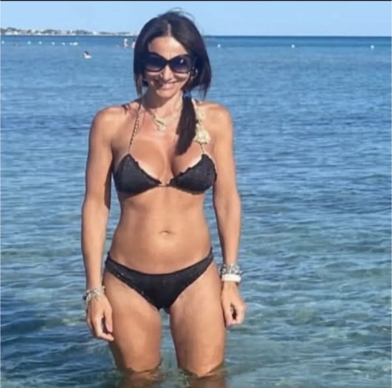 Eine Frau im schwarzen Bikini steht im flachen Meer. Sie trägt Sonnenbrille, Armbänder und hat dunkles Haar. Hinter ihr ist das Meer und der Horizont zu sehen.