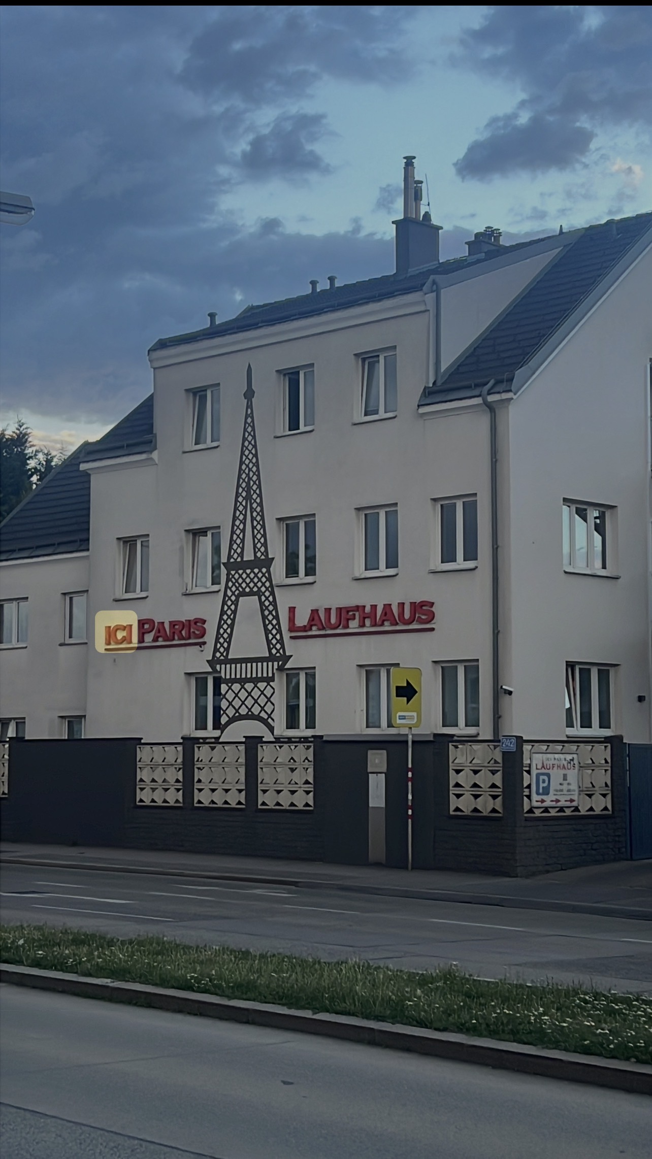 Ein Gebäude mit laubhaus-Schriftzug, Eiffelturmsymbol und ICI PARIS-Logo bei Dämmerung. Ansicht von der Straße, Vordergrund mit Rasen.