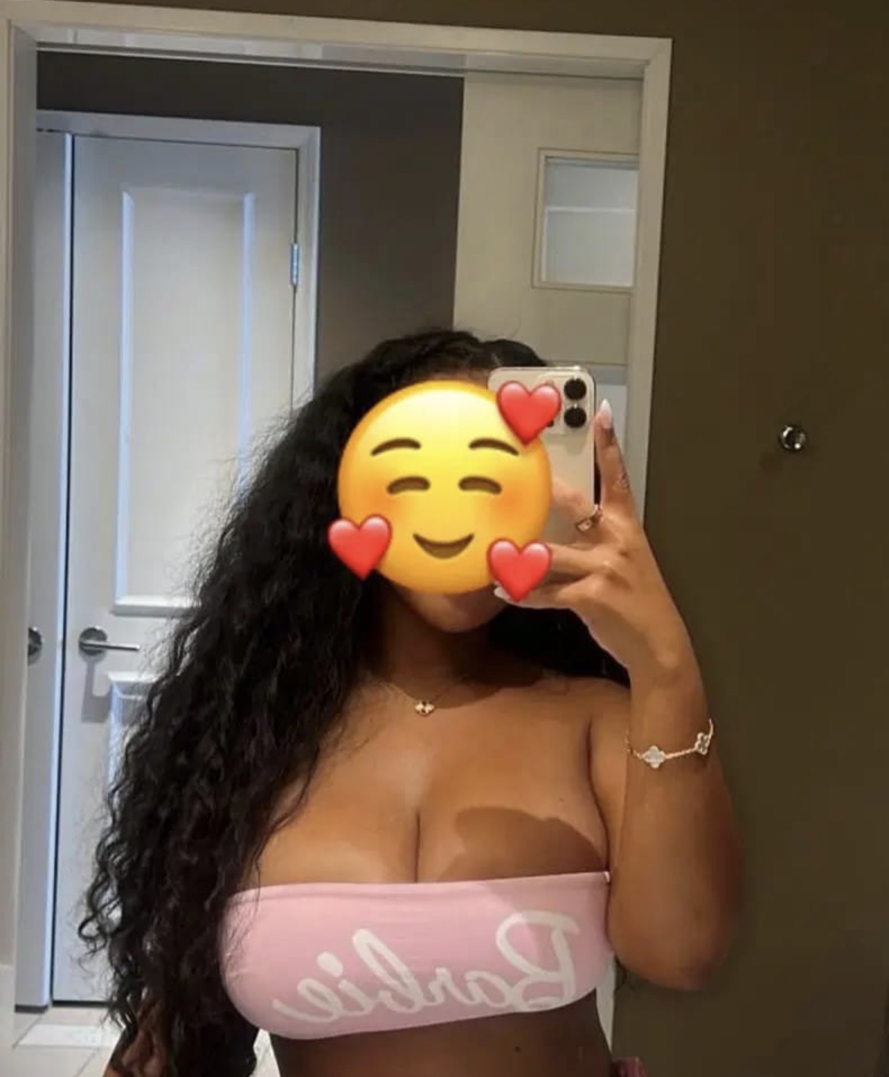 Eine Frau mit langen Locken, rosafarbenem Bustier und einem Emoji-Verschleier auf dem Gesicht macht ein Selfie im Spiegel. Die Szene spielt in einem Badezimmer.