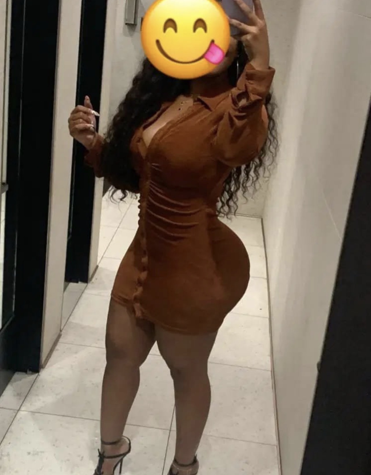 Eine Person mit dunklem, lockigen Haar trägt ein bodenlanges, braunes Kleid und posiert vor einem Spiegel. Sie hält eine Zigarette in der Hand und trägt Stilettos. 🍌