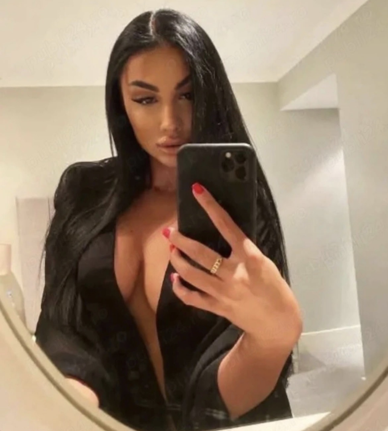 Eine Frau mit langen schwarzen Haaren und einem tief ausgeschnittenen schwarzen Blazer macht im Badezimmerspiegel ein Selfie mit einem Smartphone.