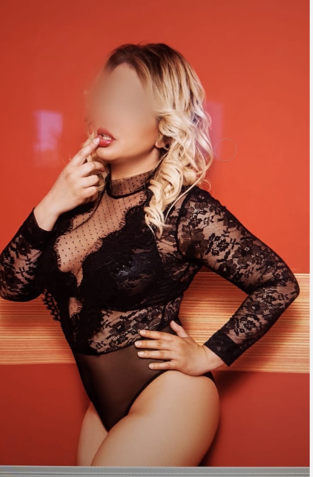 Eine Person mit blonden Locken sitzt auf einer Holzbank vor rotem Hintergrund. Sie trägt ein schwarzes Spitzenoberteil und hält die Hand an den Mund.
