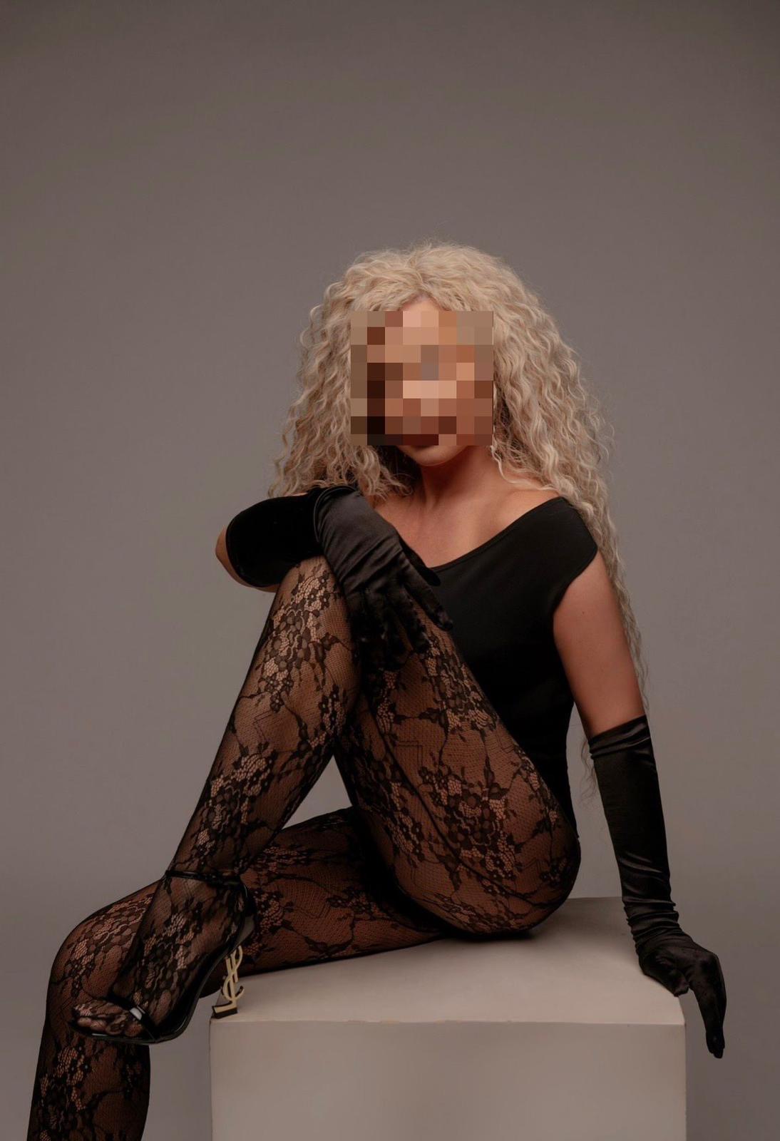 Eine weibliche Person mit blonden Locken trägt schwarze Spitzenstrümpfe, Handschuhe und sitzt auf einem Podest.