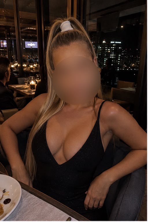 Eine Frau mit langen blonden Haaren in einem tief ausgeschnittenen schwarz glänzenden Kleid sitzt an einem Tisch in einer stilvollen Bar mit nächtlicher Skyline im Hintergrund. Gesicht unscharf.