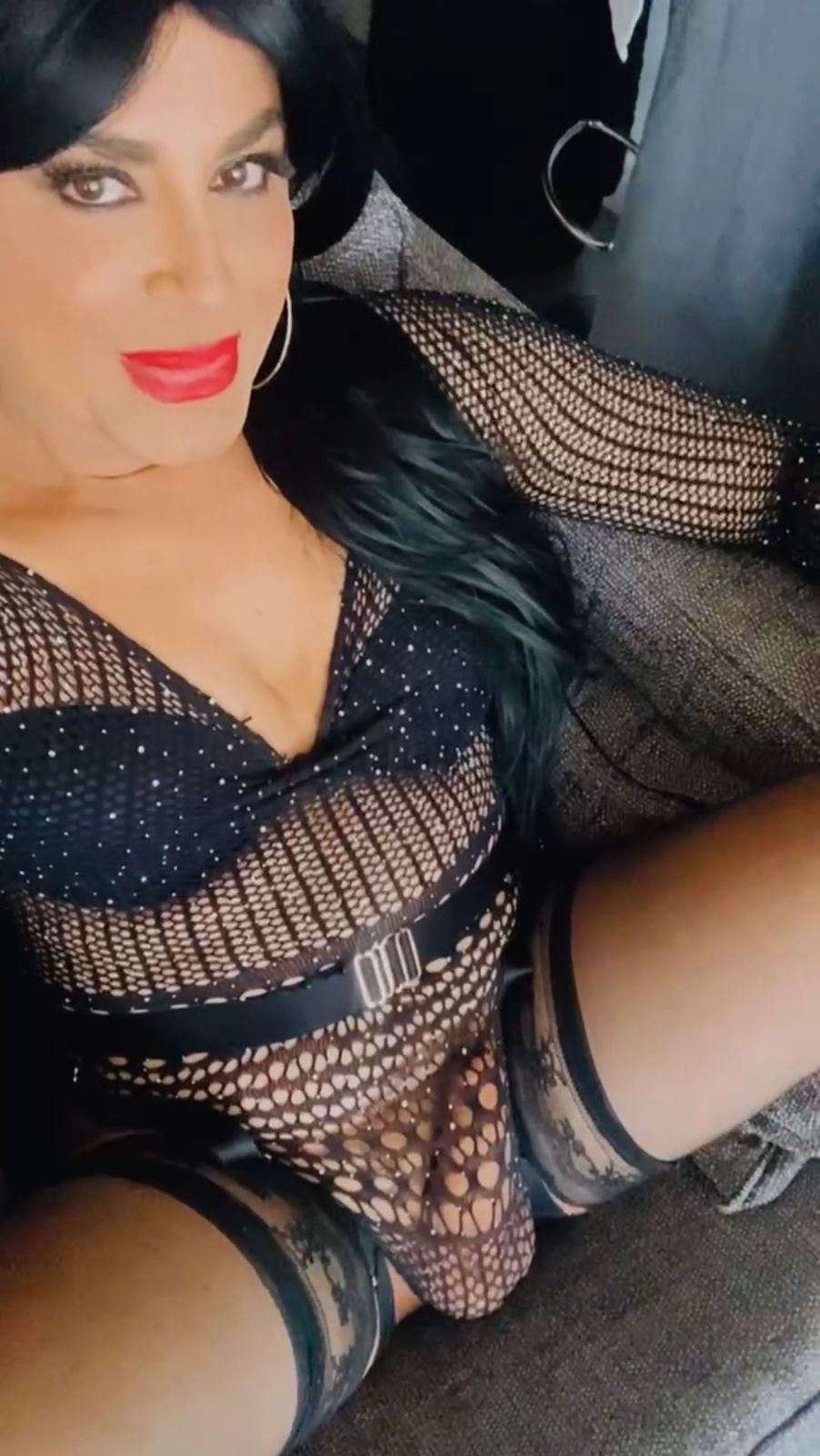Eine Frau mit dunklem Haar trägt ein teddyähnliches, schwarzes Netzkleid mit Pailletten und Reißverschluss an der Seite. Ihre Pose ist elegant.