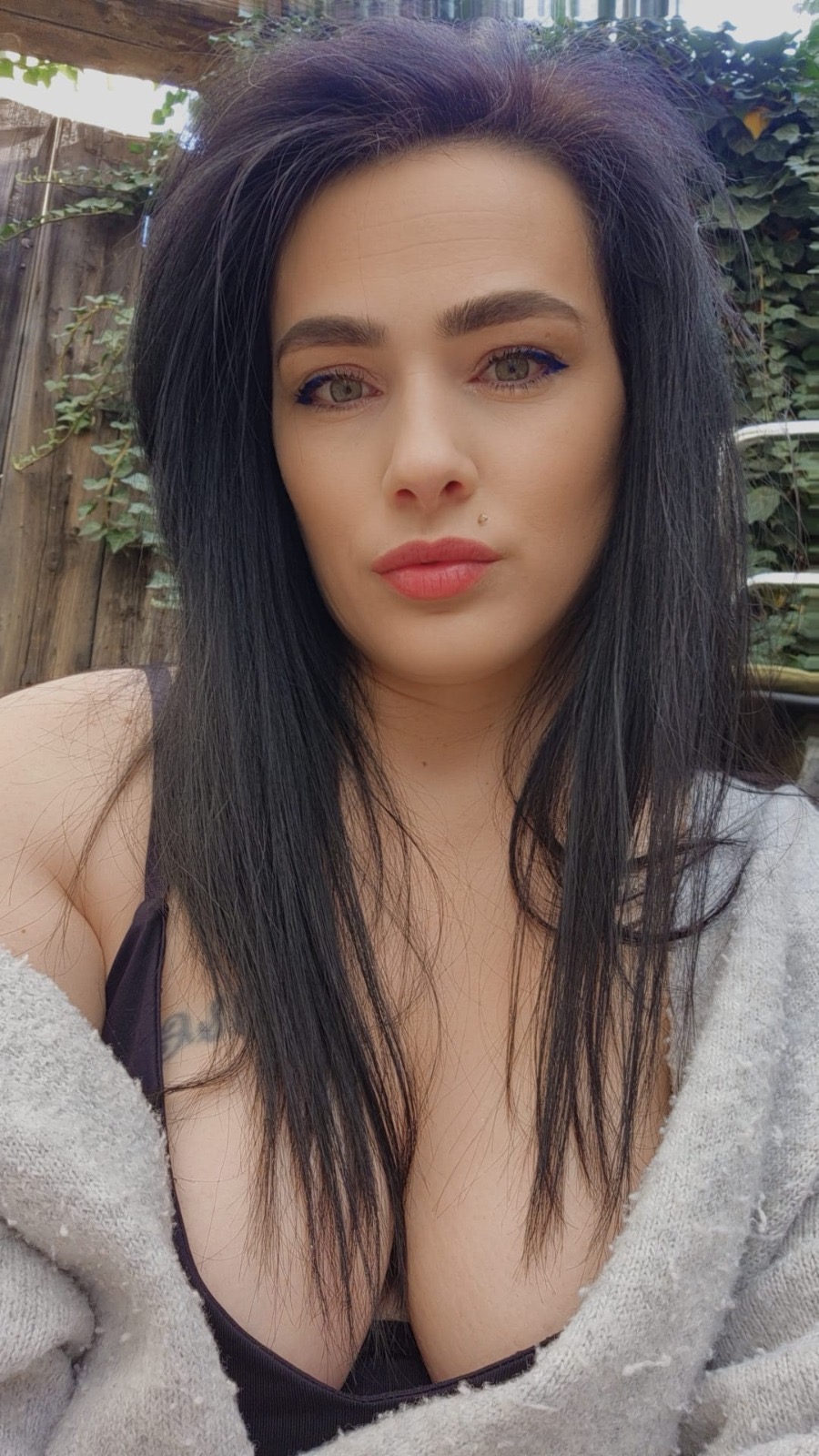 Eine Frau mit langen schwarzen Haaren und grßnen Augen trägt ein dunkles Top und einen grauen Pullover. Hinter ihr sind Holzzäune und Pflanzen zu sehen.