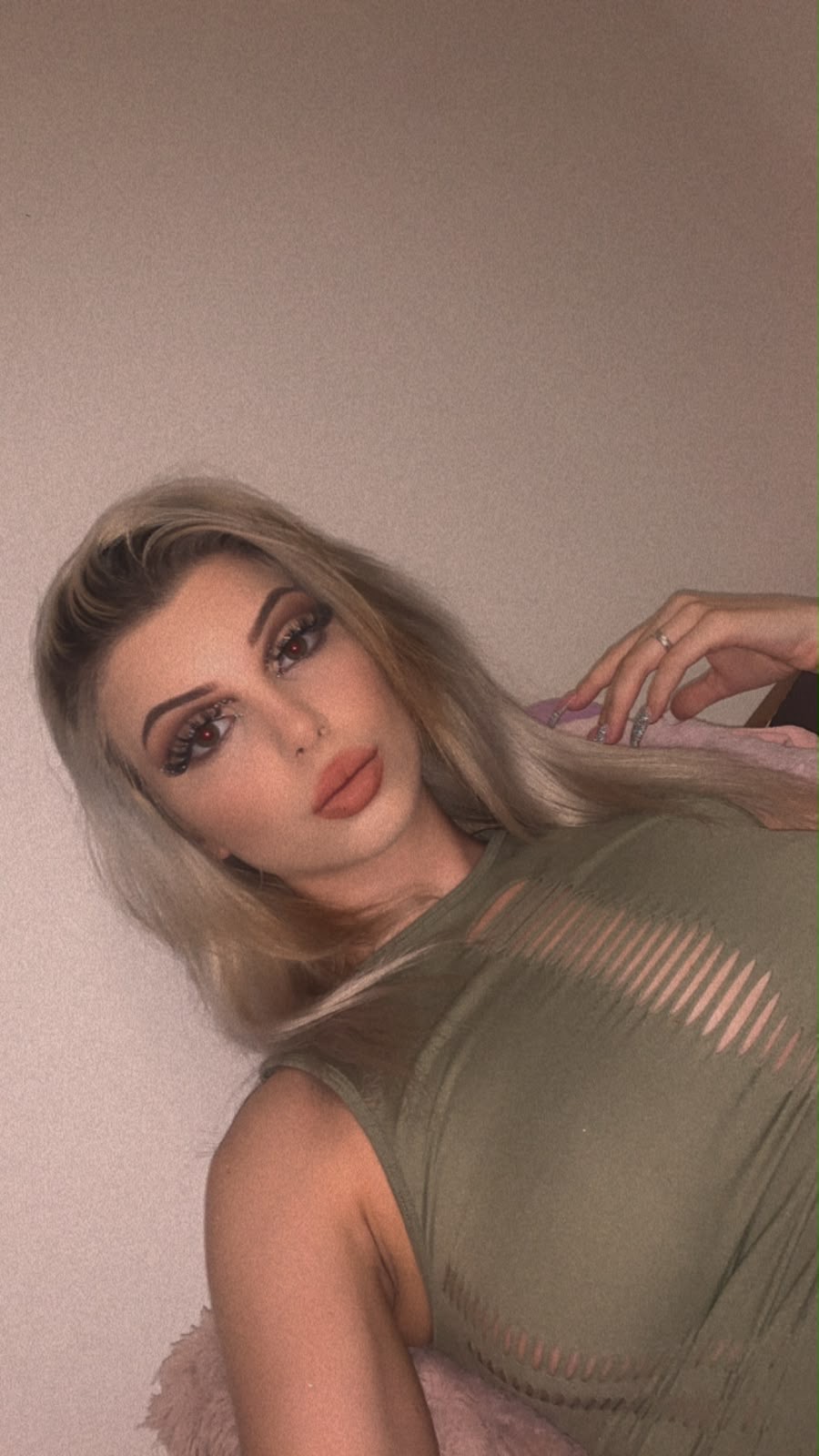Eine Frau mit blonden Haaren und sorgfältig gestalteter Varieté-Schminke liegt durch einen Schlitz in einem olivgrünen Kleid. Ein Siegelring am Mittelfinger ihrer rechten Hand. Lila:Lila.