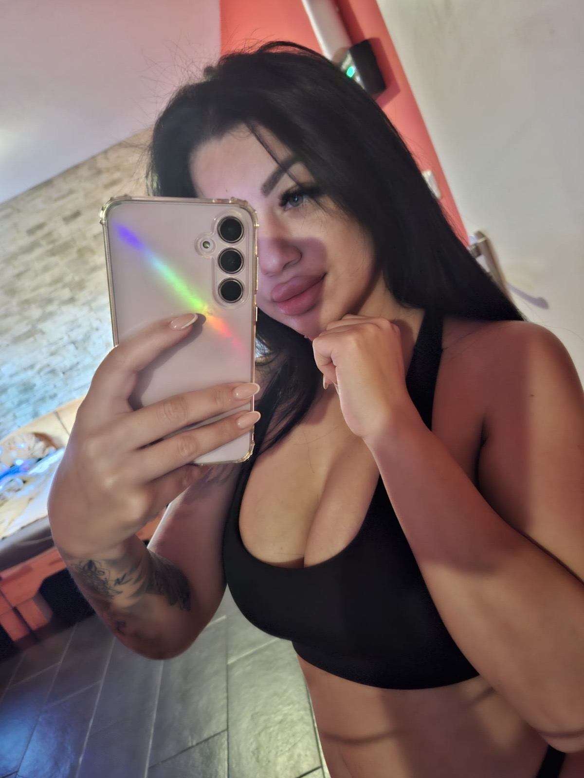 Eine Frau mit dunklen Haaren und Tattoo am Unterarm hält ein Smartphone vor ihr Gesicht und posiert im Spiegel in einem schwarzen Top.