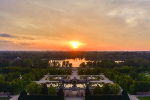 Sonnenuntergang über einem Schlosspark mit Spiegelpool, geometrischen Gärten und einem See im Hintergrund.
