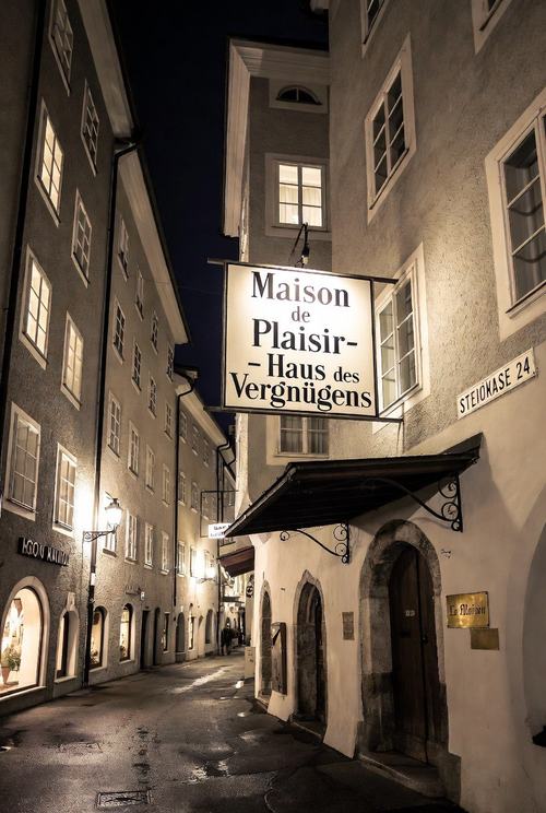 Nachtstraße mit beleuchteten Gebäuden, Schild "Maison de Plaisir - Haus des Vergnügens" und Straßenschild "STEIGKASE 24".