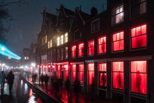 Nachtansicht einer Straße mit roten beleuchteten Fenstern, Menschen gehen entlang eines Kanals, typisch für das Rotlichtviertel in Amsterdam.
