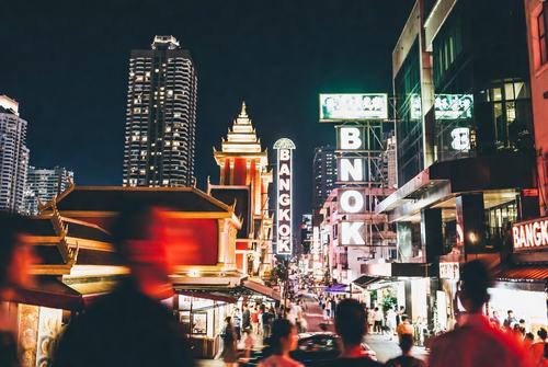 Nächtliche Straßenszene in Bangkok mit beleuchteten Gebäuden, Menschenmenge und großem "Bangkok"-Schild im Vordergrund.