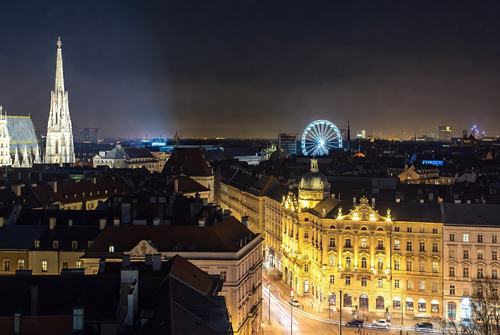 Nachtstadt mit beleuchtetem Kirchturm, historischen Gebäuden und einem großen, leuchtenden Riesenrad im Hintergrund.