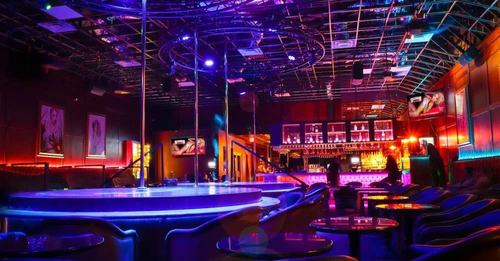 Bunte Bar mit Tanzfläche, Pole-Dance-Stangen, leuchtenden Möbeln und beleuchteter Theke im Hintergrund.