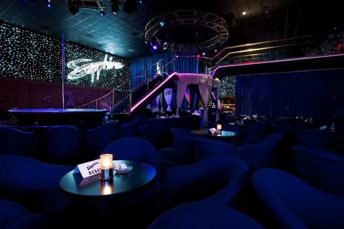 Dunkle Lounge mit blauen Sitzgruppen, runden Tischen, Kerzenlichtern und einer beleuchteten Treppe im Hintergrund.