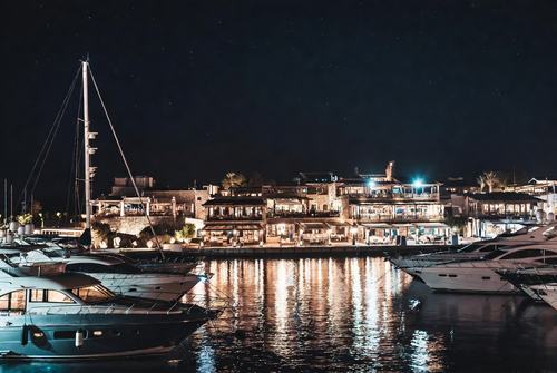 Nachtansicht eines beleuchteten Hafens mit Yachten und Häusern im Hintergrund.