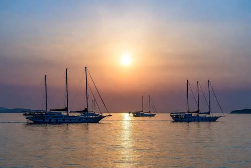 Drei Segelschiffe bei Sonnenuntergang auf ruhigem Meer, Reflexion des goldenen Lichts im Wasser.